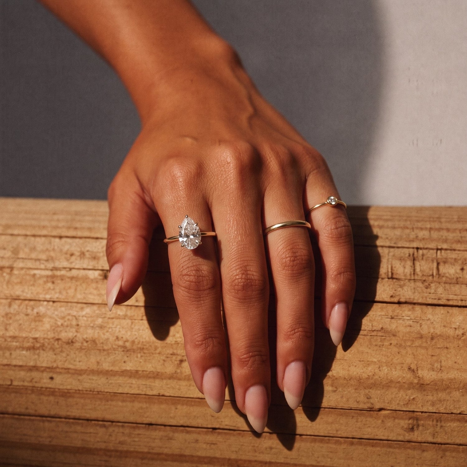 Three gold diamond rings on a hand resting on timber, a pear solitaire, a plain band and a bezel band