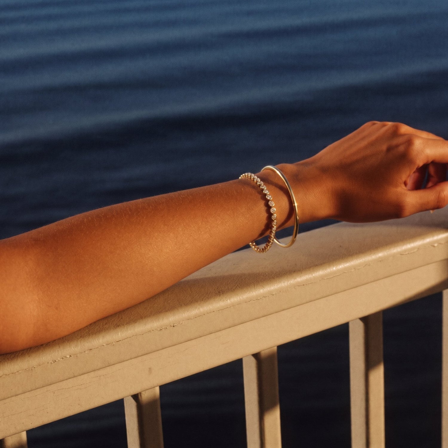 Gold diamond line bracelet and plain gold bangle on a forearm resting on a balcony rail above the ocean