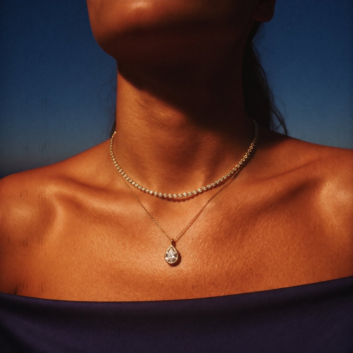 Gold diamond line necklace layered with a pear diamond pendant against a dusk sky