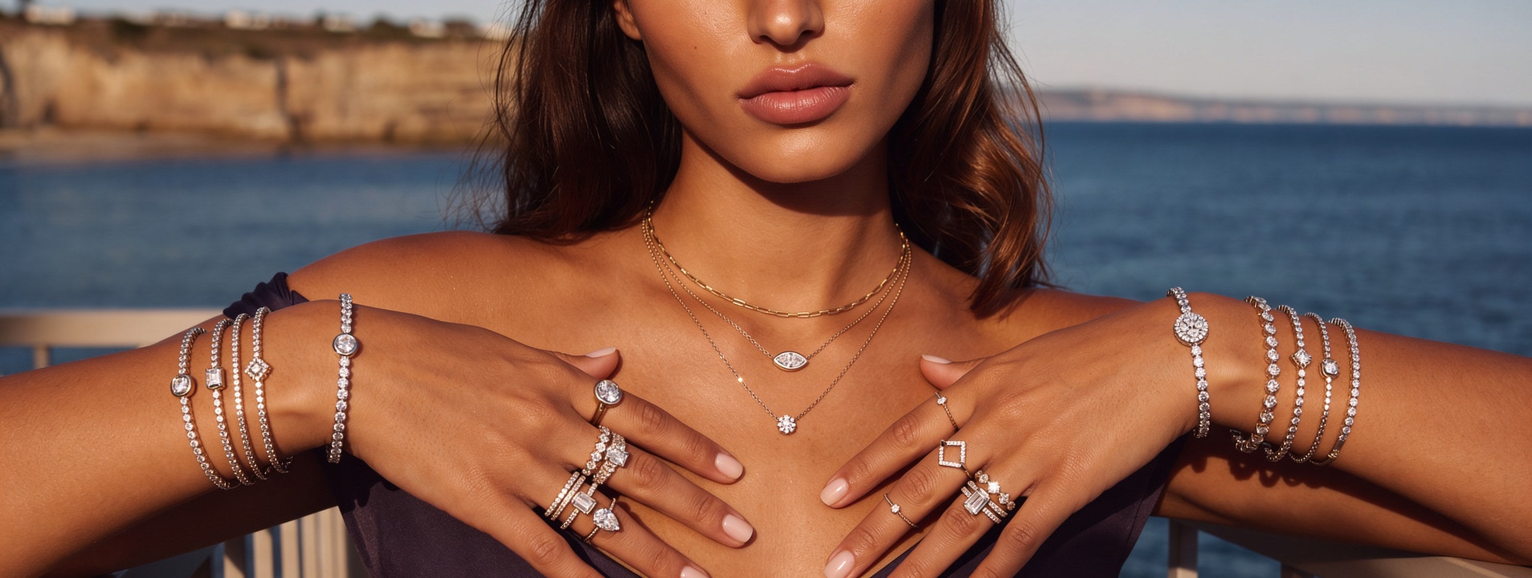Woman on a clifftop balcony above the ocean wearing stacked Divara diamond tennis bracelets, layered necklaces and rings