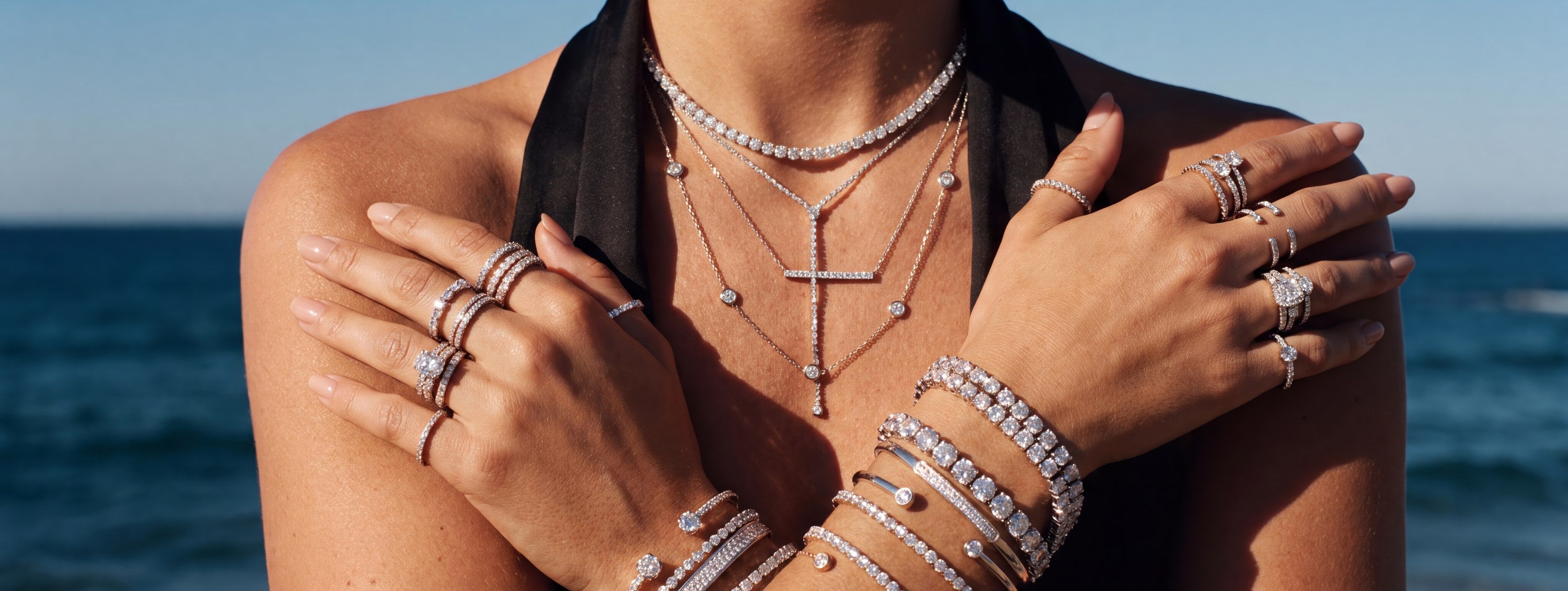 Crossed arms against deep blue ocean showing layered Divara diamond necklaces, stacked tennis bracelets and eternity rings