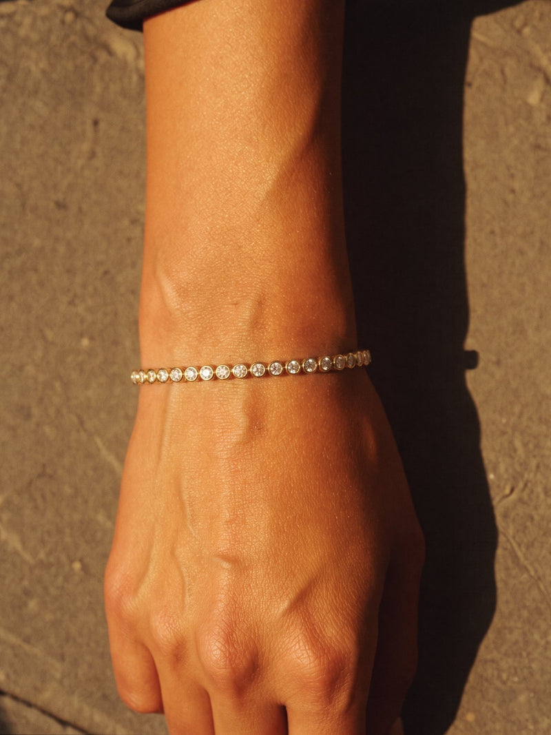 Gold bezel-set diamond line bracelet on a wrist resting on sandstone in hard afternoon sun